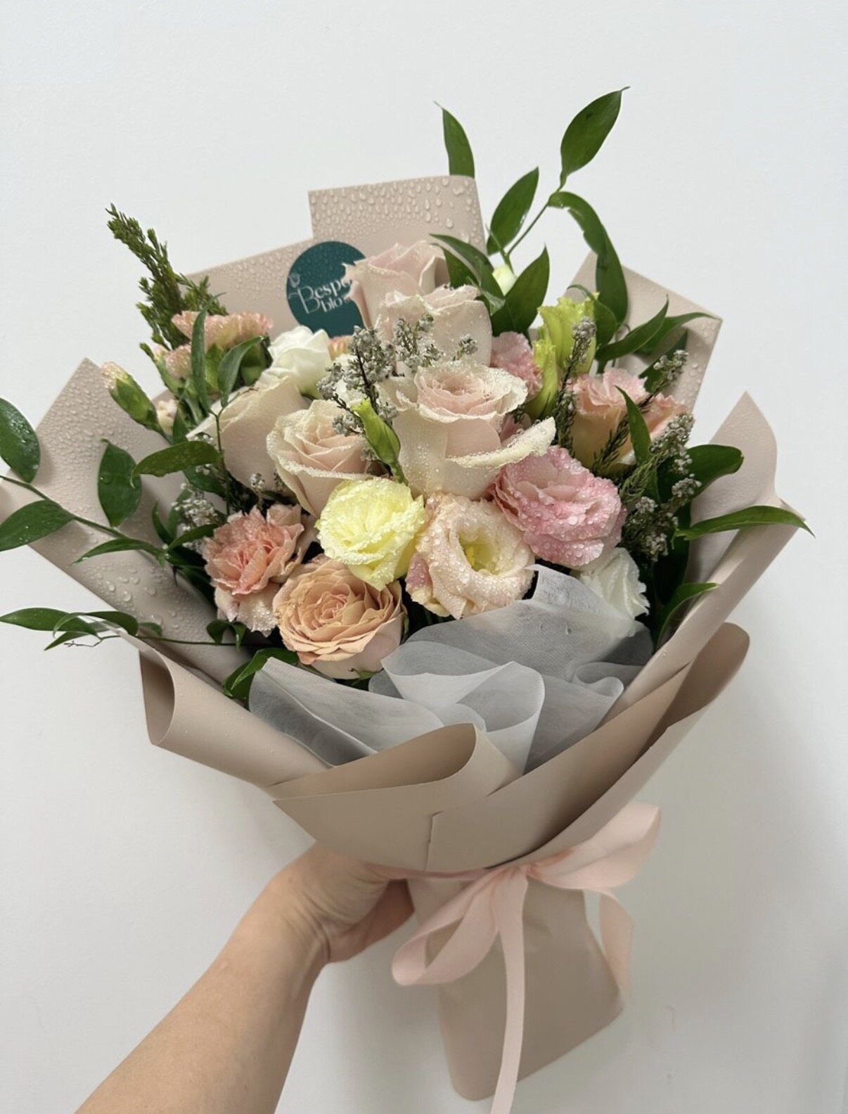A hand-tied bouquet of blush and cream roses with pink lisianthus and peach spray carnations, wrapped in taupe paper with a pink ribbon.