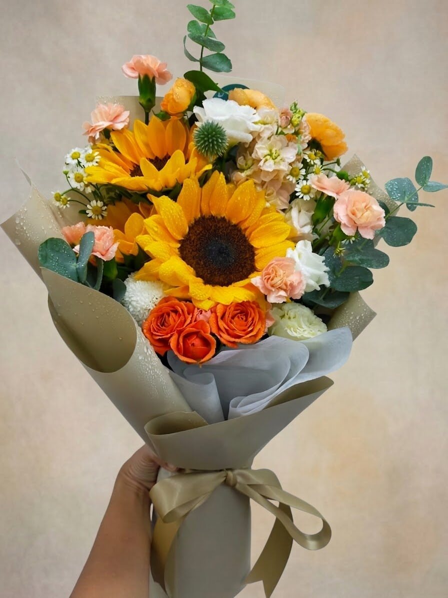 A hand-tied bouquet of yellow sunflowers with orange spray roses, peach carnations and white chamomile, wrapped in olive-green paper.